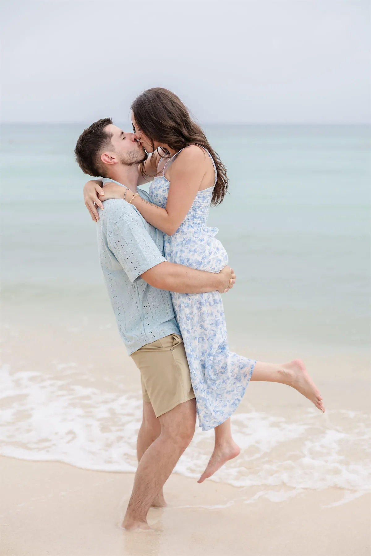 A man lifting a woman in a blue floral dress for a kiss on the shoreline in Cozumel.