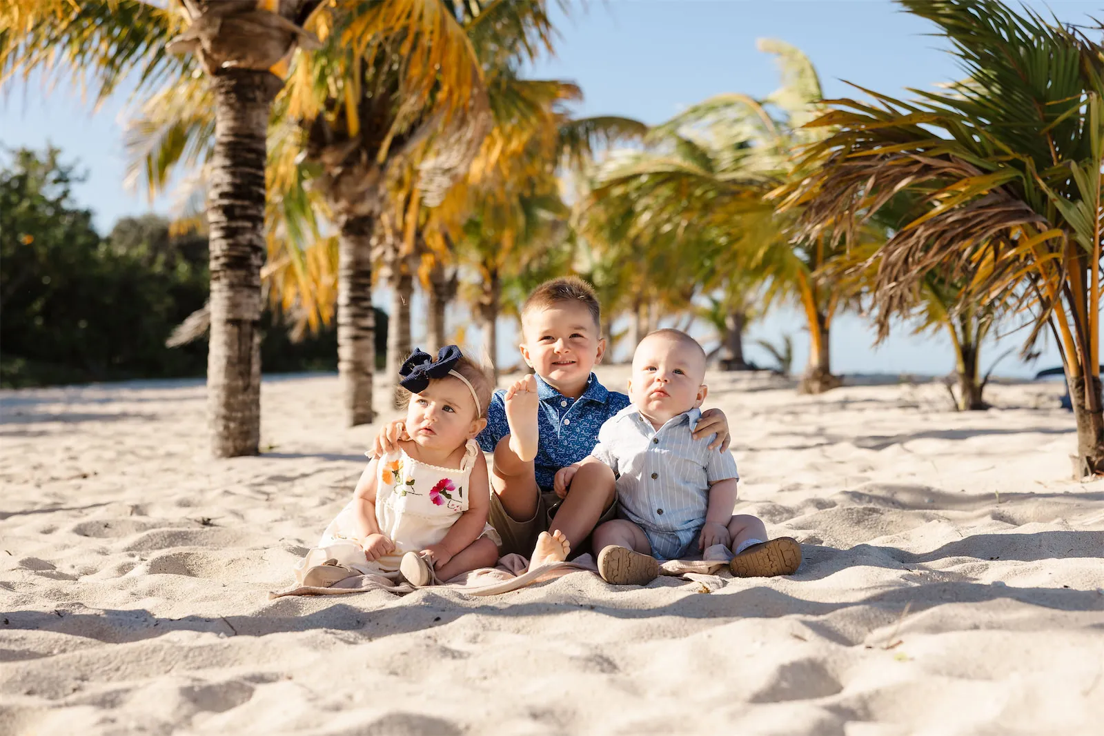 Three young children sitting and playing together on the sand under palm trees during a family photo session in Cozumel.