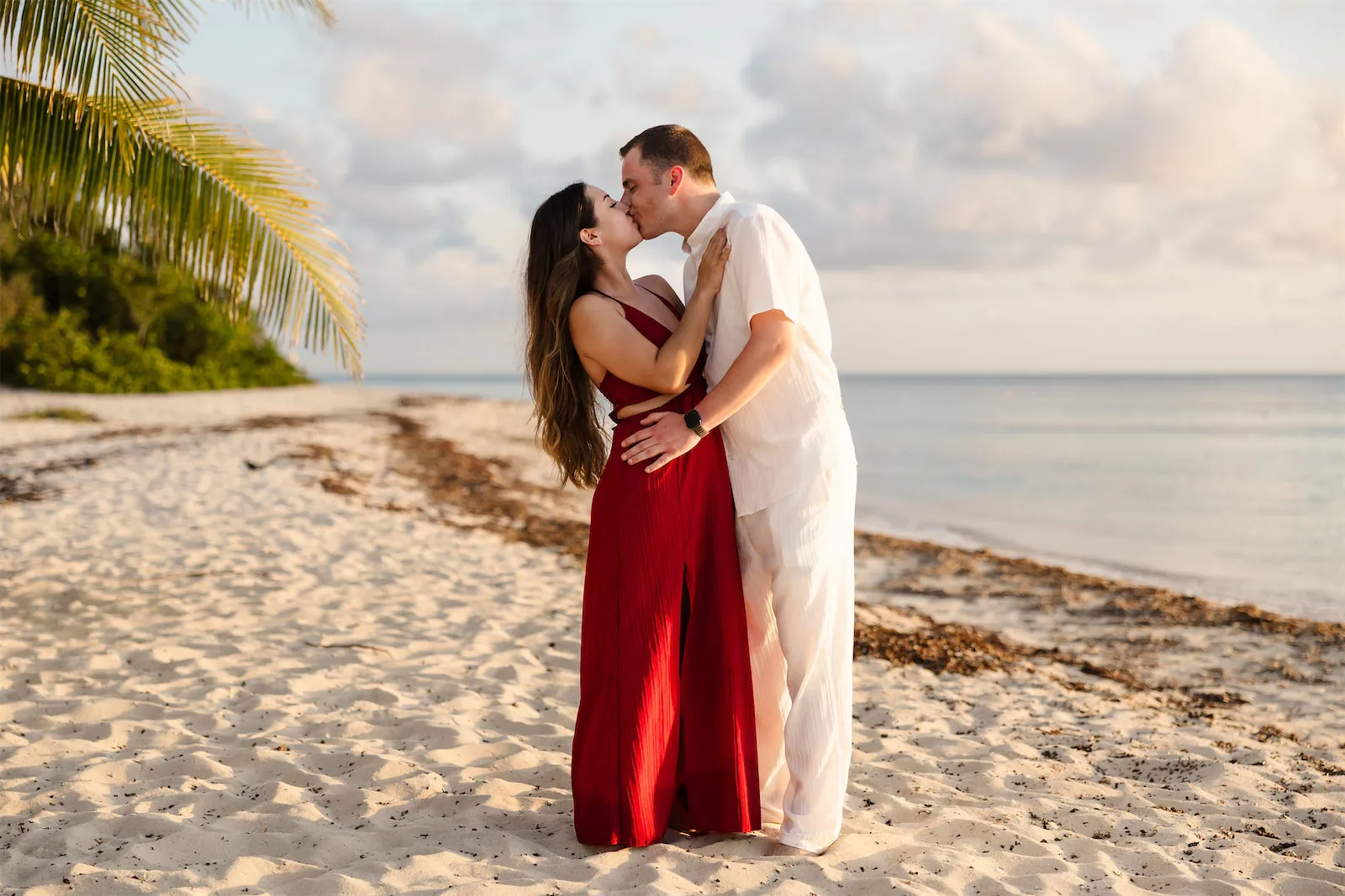 A couple in white and red outfits kissing on the beach during a romantic golden hour photo shoot.
