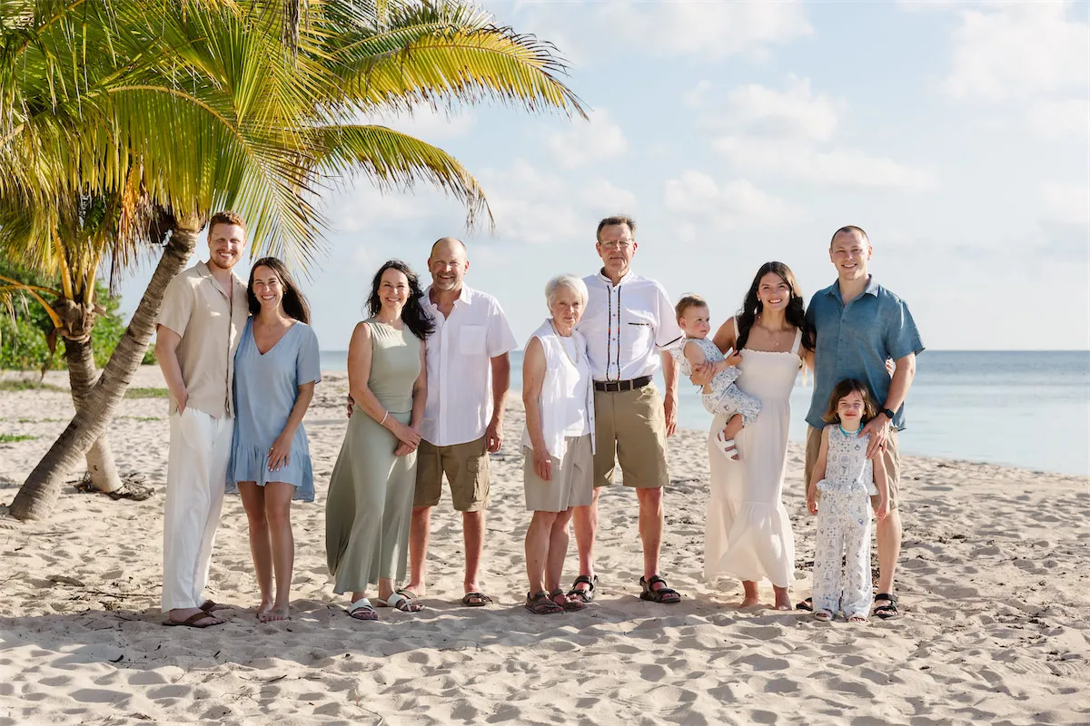 A large extended family of ten posing for a sunny beach portrait with a leaning palm tree and blue ocean in Cozumel.