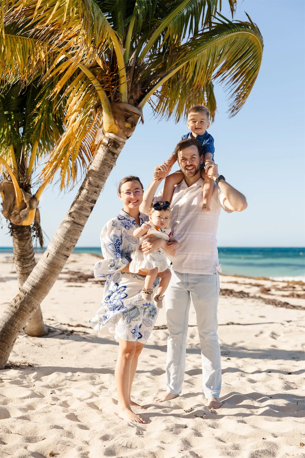 A happy family of four with a toddler on his father's shoulders posing by a palm tree on a Cozumel beach.
