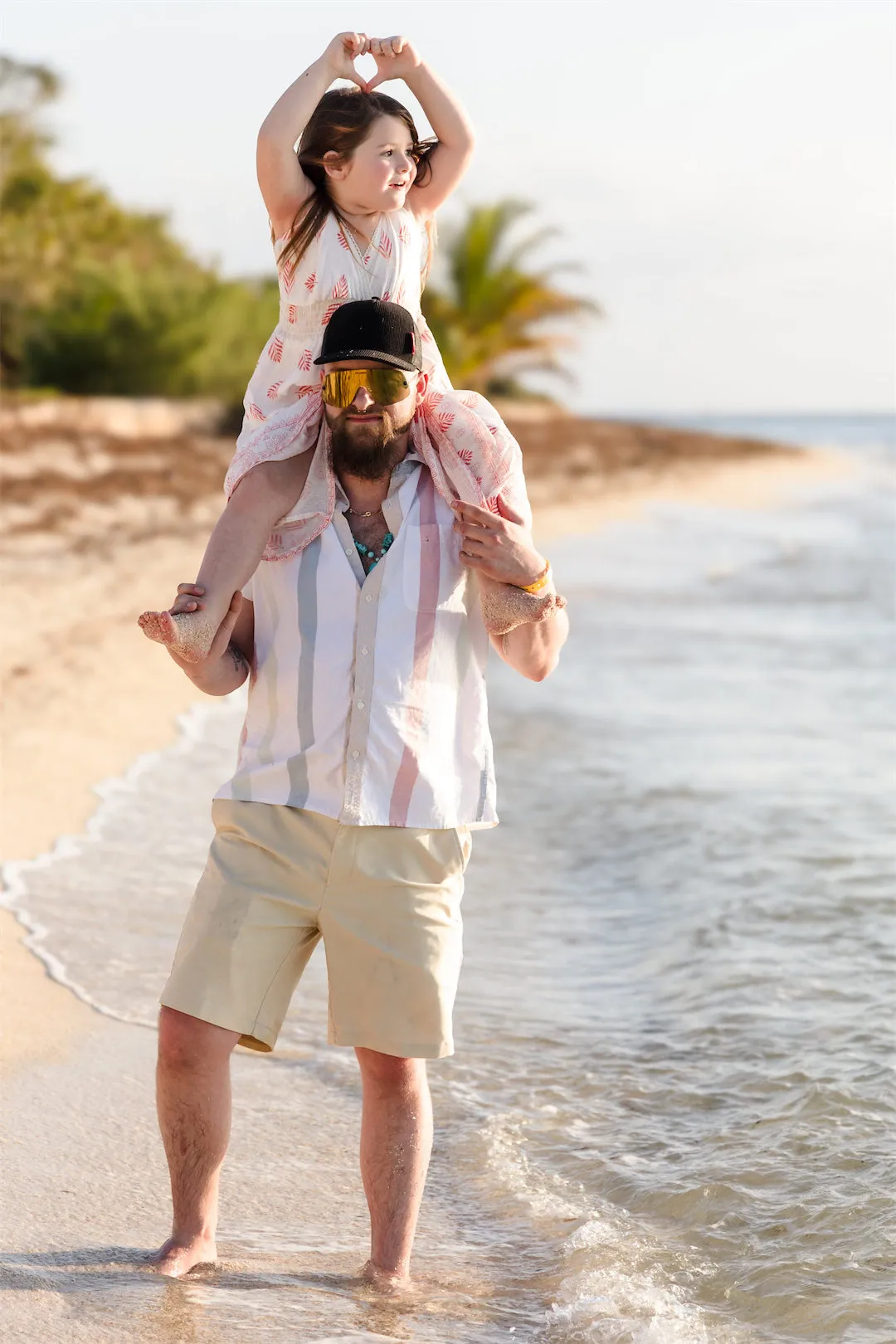 A father carrying his daughter on his shoulders while she makes a heart shape with her hands on a Cozumel beach.