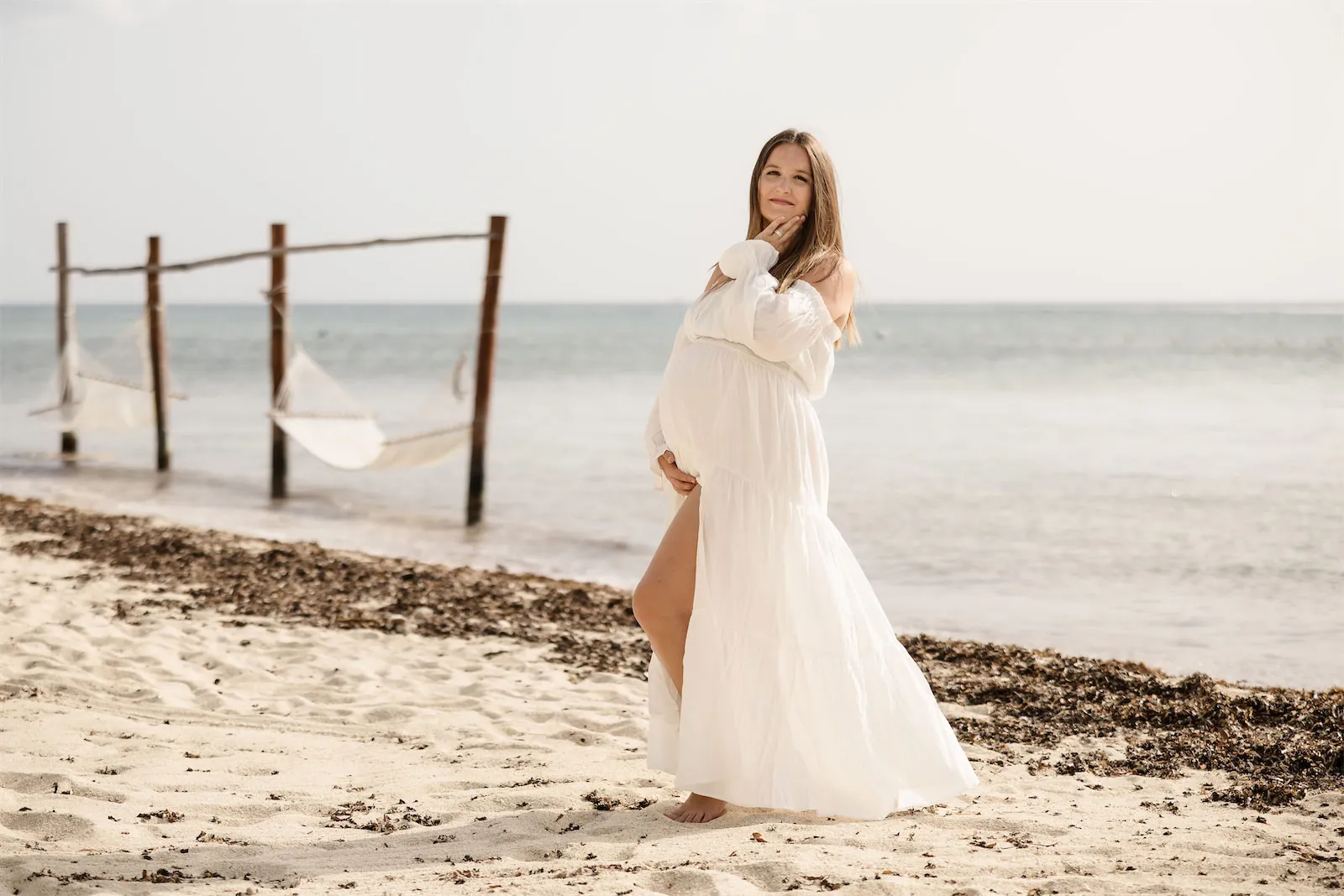 A beautiful maternity portrait of a woman in a white dress standing on the beach at Dreams Natura with ocean hammocks in the background.