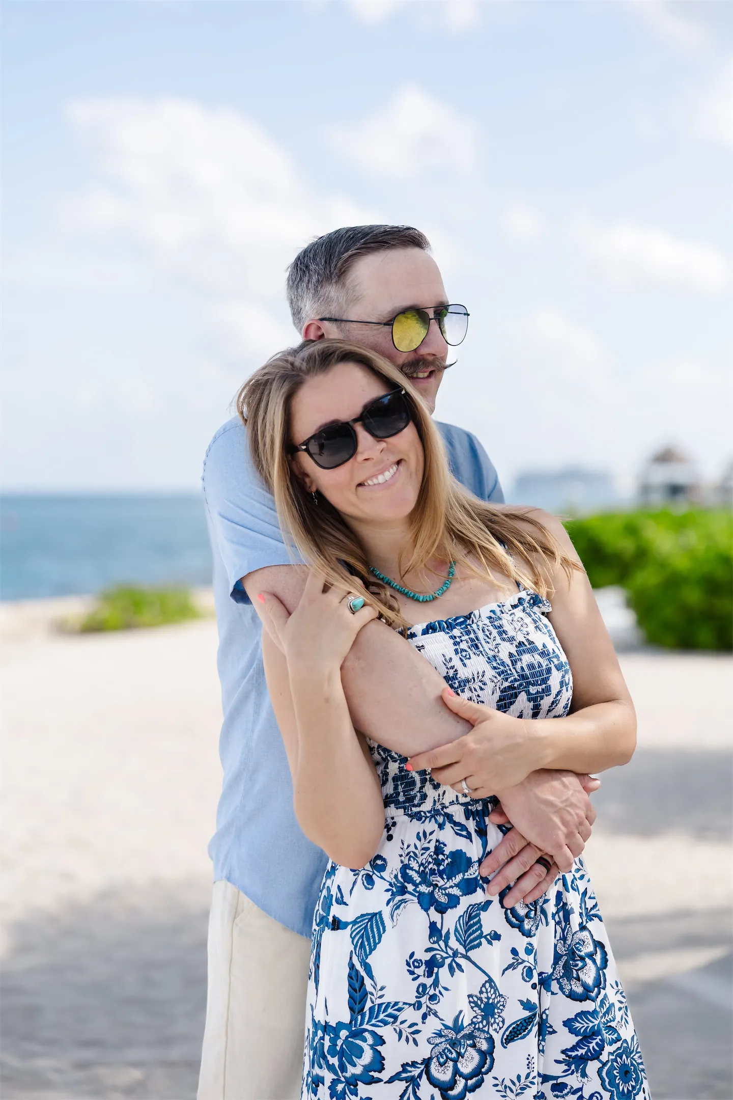 A man embracing a woman from behind as they smile on a white sand beach in Cozumel.
