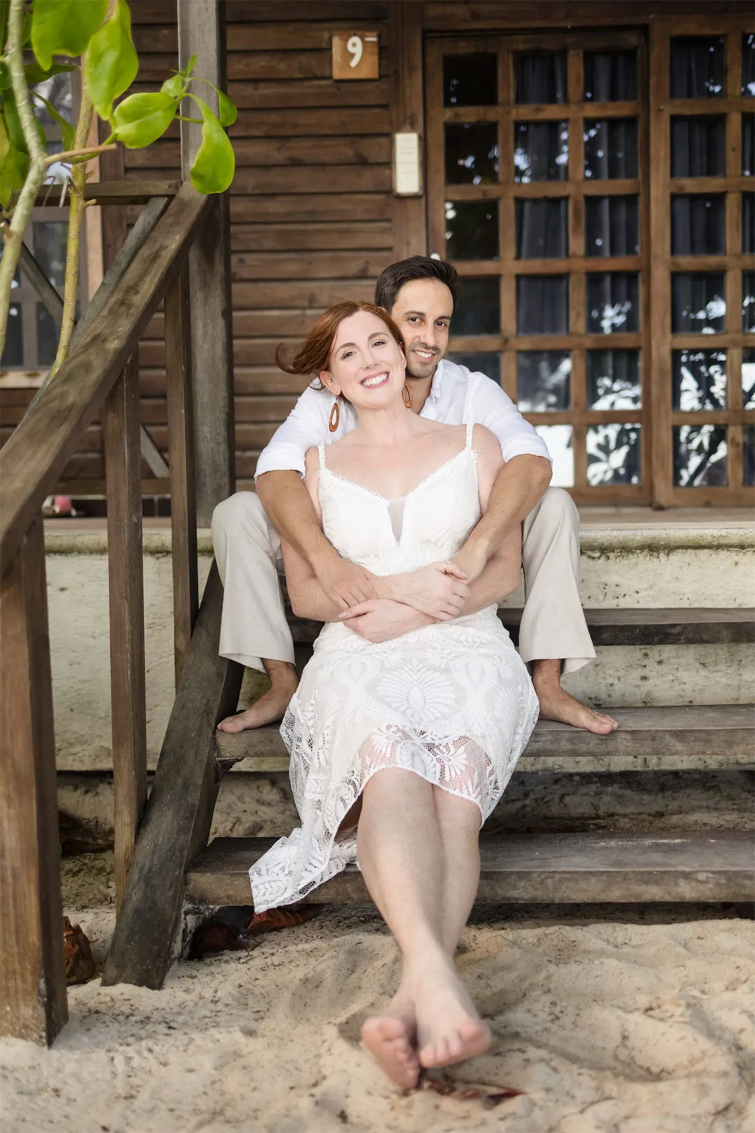 A smiling couple sitting together on the wooden steps of a tropical beach bungalow in Cozumel.