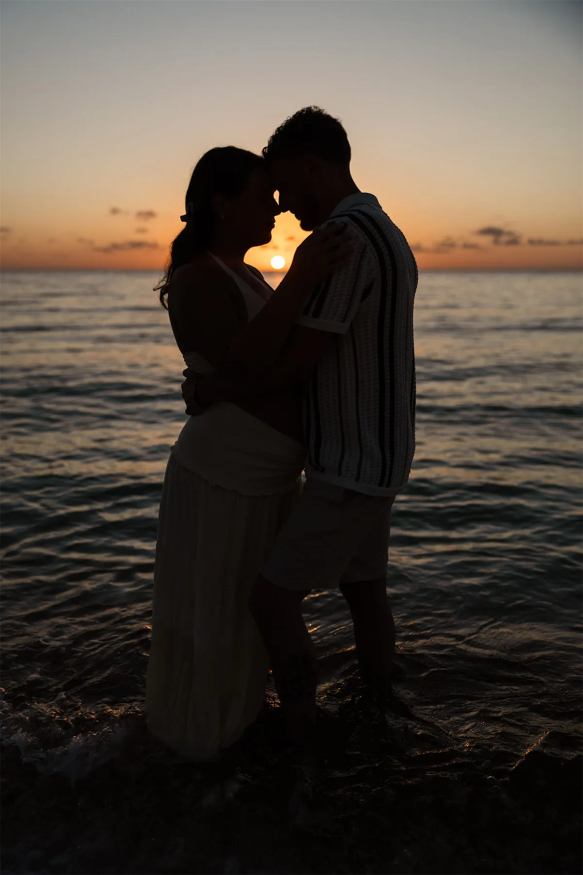 A sunset silhouette of a couple embracing in the water during a Cozumel maternity photo shoot.