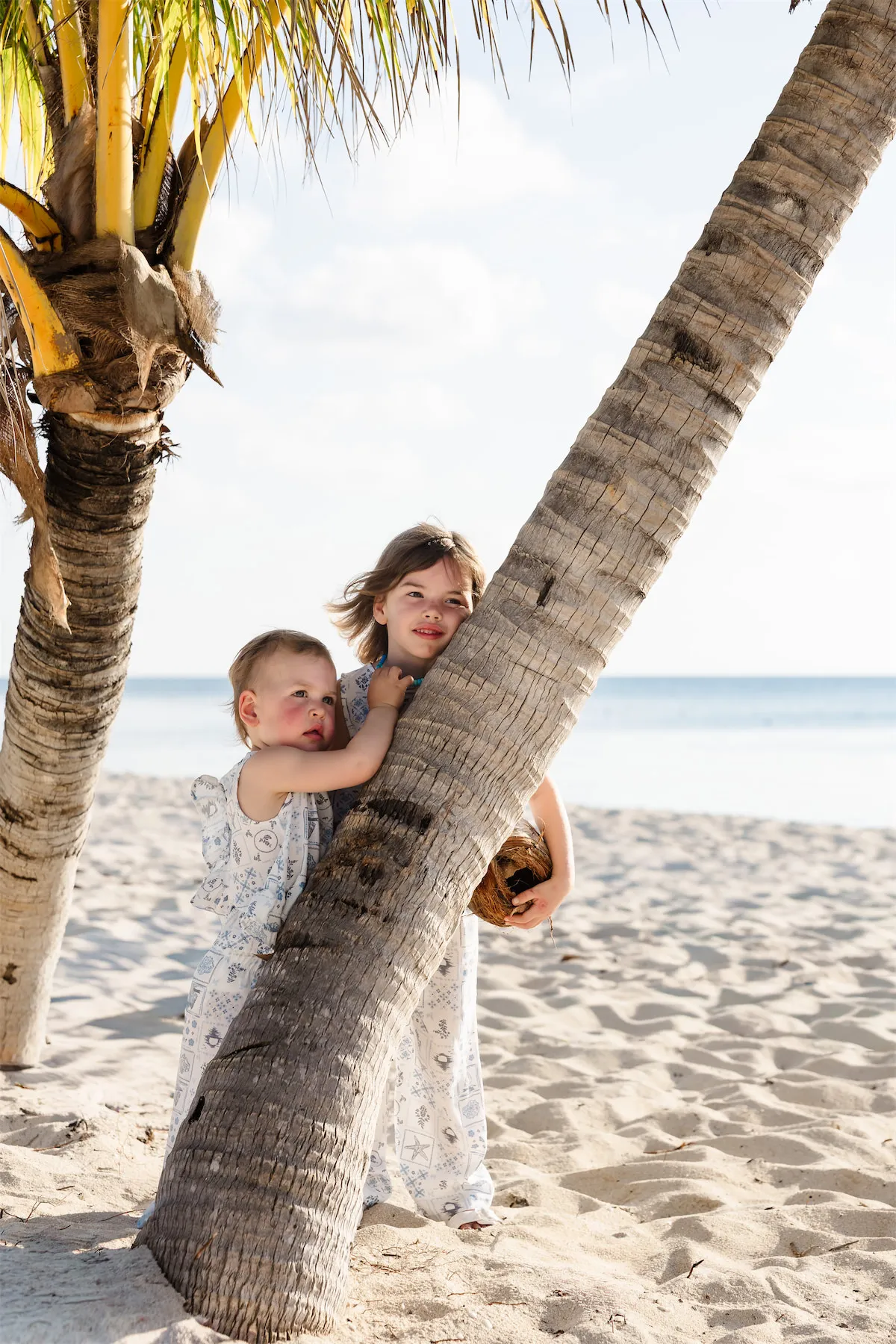 Two young sisters in matching blue and white outfits playing by a leaning palm tree during a Cozumel family photo session.